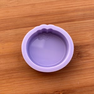 Puede incluir: Un molde de silicona circular de color lavanda con un borde decorativo. El molde tiene un interior liso y brillante y está diseñado para la artesanía. El molde está sobre una superficie de madera.