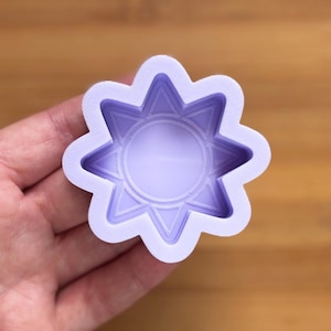 Puede incluir: Un molde en forma de estrella de color lila claro con ocho puntas redondeadas. El centro tiene una hendidura circular y las puntas de la estrella tienen detalles triangulares. El molde se sostiene en una mano, con una superficie de madera en el fondo.