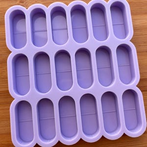Könnte beinhalten: Eine lavendelfarbene Silikonform mit 15 pillenförmigen Hohlräumen. Jeder Hohlraum hat eine Mittellinie, und die Form ist auf einer Holzoberfläche platziert. Die Form ist für das Basteln oder Erstellen kleiner, gleichmäßiger Formen konzipiert.