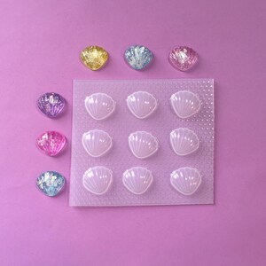 Könnte beinhalten: Eine transparente Silikonform mit neun muschelförmigen Vertiefungen. Die Form befindet sich auf einer violetten Oberfläche. Es gibt neun schillernde Muscheln, einige rosa, einige blau und einige gelb, die um die Form herum verstreut sind.