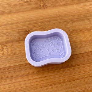 Puede incluir: Un molde de silicona de color lila claro con forma rectangular y bordes redondeados. El molde presenta un interior texturizado con círculos de varios tamaños, creando un patrón de burbujas. El molde está diseñado para manualidades y proyectos de bricolaje.