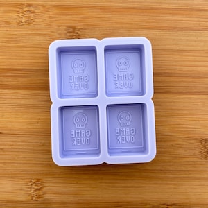 Pode incluir: Um molde de silicone roxo com quatro cavidades quadradas. Cada cavidade tem um gráfico de caveira e o texto "GAME OVER" inscrito dentro.
