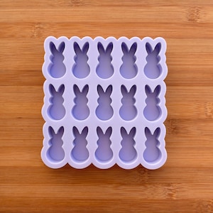 Può includere: Uno stampo in silicone lilla chiaro con bordo smerlato, con 15 cavità a forma di coniglietto. Lo stampo è progettato per creare dolcetti a tema pasquale, come cioccolatini o caramelle. Lo stampo è posto su uno sfondo di legno.