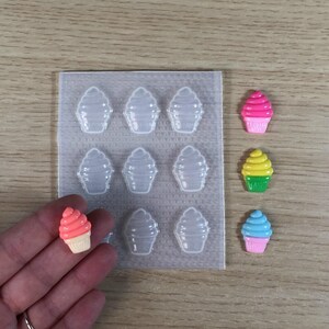 Puede incluir: Un molde de plástico transparente con nueve cavidades en forma de cupcake. Hay cuatro amuletos de cupcake terminados en diferentes colores: rosa, amarillo, verde y azul.