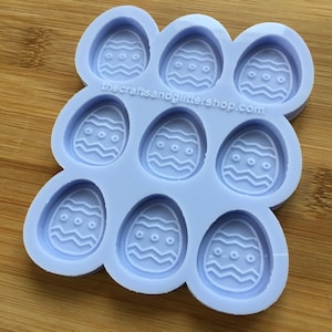 Peut inclure: Un moule en silicone de couleur violet clair avec neuf cavités en forme d'œufs de Pâques. Chaque œuf a un motif de lignes ondulées et trois petits cercles.