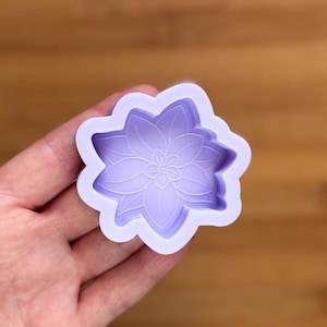 Puede incluir: Un molde de silicona de color lavanda con forma de flor. El molde tiene un diseño floral detallado con múltiples capas y un borde festoneado. El molde se sostiene en una mano sobre un fondo de madera borroso.