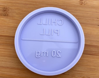 May include: A circular light purple silicone mold featuring embossed text that reads CHILL PILL with a horizontal line across the center and the text 20 mg below it. The mold has a raised outer rim and a smooth, flat interior surface for casting.