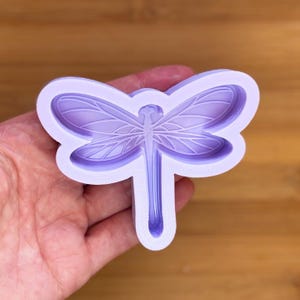 Puede incluir: Un molde en forma de libélula de color lavanda. El molde tiene patrones detallados de alas y cuerpo. La libélula se sostiene en una mano sobre un fondo de madera. Este molde es probablemente para manualidades o repostería.