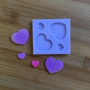 Puede incluir: Un molde cuadrado de silicona de color lila claro con cavidades en forma de corazón. Varias piezas de resina en forma de corazón de diferentes tamaños y colores, incluyendo púrpura y rosa brillantes, están esparcidas alrededor del molde sobre una superficie de madera. Los corazones son probablemente para manualidades.