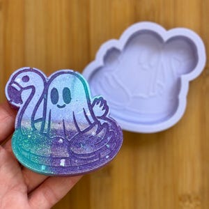 May include: A handmade resin ghost-shaped charm floating on a flamingo floatie, with glitter accents in shades of purple, teal, and blue. A matching silicone mold is next to the charm.