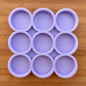 Può includere: Uno stampo in silicone color lavanda con nove cavità circolari. Lo stampo è progettato per la creazione di piccoli oggetti rotondi. Le cavità sono uniformemente distanziate e hanno una finitura liscia e lucida. Lo stampo è posto su una superficie di legno.