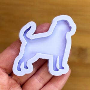Peut inclure: Un moule en silicone violet clair en forme de chien. Le moule est tenu dans une main et a une finition lisse et brillante. La silhouette du chien est détaillée, montrant le contour de l'animal.