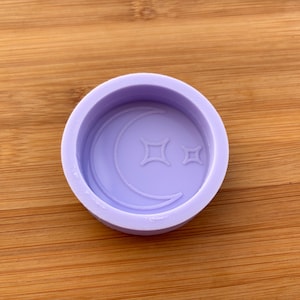 Puede incluir: Un molde redondo de color lavanda con una luna creciente y dos formas de estrella. El molde está diseñado para la artesanía y la creación de elementos decorativos. El diseño está en relieve dentro de la forma circular.
