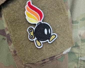Op de afbeelding: Een geborduurde patch met een zwart bolvormig figuur met een wit oog, gele voeten en een gele opwindsleutel aan de zijkant. Een vlam met rode, oranje en gele gradiënten steekt uit de bovenkant van de bol. De patch is bevestigd op een beige stof.