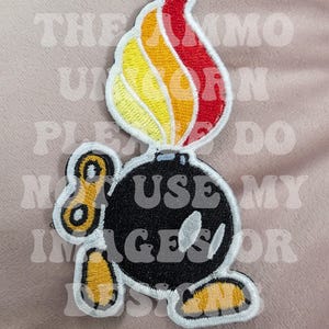 May include: Embroidered patch featuring a cartoon bomb with a flame. The bomb is black with white accents and the flame is red, orange, and yellow. The patch has a white border and a key-like design.