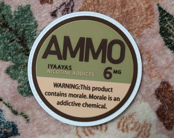 Op de afbeelding: Een ronde sticker met het woord "AMMO" in donkerbruine letters. Daaronder staat "IYAAYAS NICOTINE ADDICTS" en "6 MG". De onderste sectie bevat een waarschuwing over het product dat moraal bevat, een verslavende chemische stof.