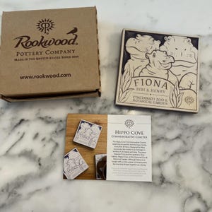 May include: A square ceramic coaster featuring a hippo family illustration with the text "FIONA BIBI & HENRY CINCINNATI ZOO & BOTANICAL GARDEN." The coaster is displayed with a wooden base, a box, and a card.