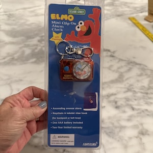 May include: A red Elmo mini clip-on alarm clock with a flashlight, packaged on a blue card. The clock face features Elmo. The package includes a keychain and lobster claw hook. The text on the package reads "Sesame Street" and "Elmo".