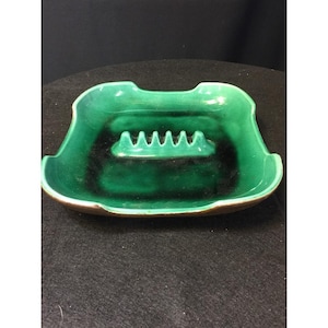 May include: A vintage green ceramic ashtray with a shield-like shape. The ashtray features a central rest area with multiple grooves for holding cigarettes. The exterior is a glossy green with a dark interior and a brown rim.