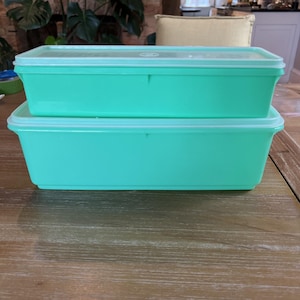 May include: Two rectangular, turquoise plastic food storage containers with clear lids are stacked on a wooden surface. The containers are empty and appear to be vintage. The lids fit snugly on top of the containers.