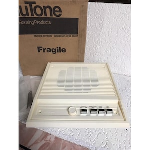 Peut inclure: Interphone NuTone blanc cassé avec grille de haut-parleur, boutons de commande et cadran. La boîte derrière indique "NuTone Housing Products" et "Fragile".