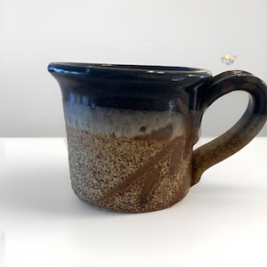 Peut inclure: Une tasse en céramique faite à la main avec un bord bleu foncé, un dégradé de gris et de marron, et une surface texturée et mouchetée. La tasse a une anse marron et des stries décoratives marron. Cette tasse unique est parfaite pour le café ou le thé.