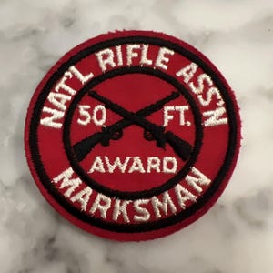 Op de afbeelding: Een ronde rode patch met wit geborduurde tekst en zwarte accenten. De tekst luidt "NAT'L RIFLE ASSN" bovenaan en "MARKSMAN AWARD" onderaan. Twee gekruiste geweren staan in het midden, met "50 FT." in het midden.