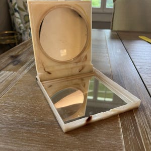 May include: Open, vintage compact mirror with a rectangular shape and a hinged lid. The compact is off-white with brown marbling. The interior features a round mirror and a square mirror. The compact is open, revealing the mirrors.