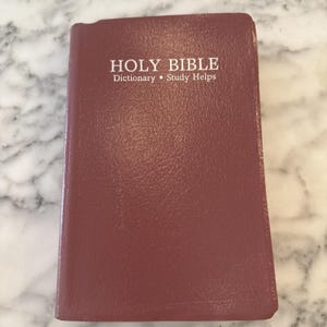 Könnte beinhalten: Eine geschlossene, weinrote Bibel mit den Worten "HOLY BIBLE" in weißer Schrift. Darunter ist der Text "Dictionary • Study Helps" zu sehen. Das Buch hat einen strukturierten Einband und liegt auf einer Marmoroberfläche.