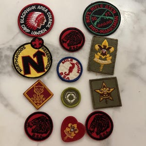 Op de afbeelding: Een verzameling vintage padvinderspatches in verschillende vormen en maten. De patches bevatten tekst, illustraties en emblemen, waaronder een Indiaans profiel, een geweer, een beer en een zwemfiguur. Kleuren zijn rood, zwart, geel, groen en wit.