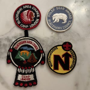 Op de afbeelding: Vier geborduurde patches met tekst en afbeeldingen. Eén patch toont een Indiaans profiel, een andere een ijsbeer, een derde een kampeerscène en de laatste een grote letter "N" met een pijl. De patches zijn in verschillende kleuren.