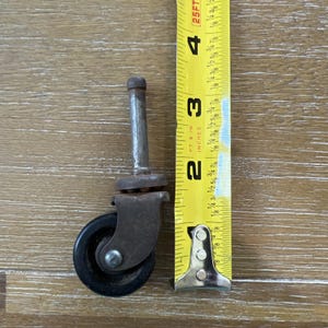 May include: A vintage furniture caster with a black wheel and metal stem, next to a yellow measuring tape. The caster is approximately 2 inches tall.