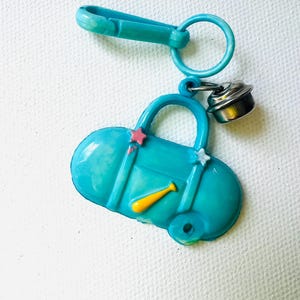 May include: A teal-colored keychain featuring a miniature duffel bag design. The bag has a yellow baseball bat, pink star accents, and a silver bell charm. The keychain includes a clasp and a ring for attachment.