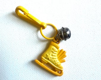 May include: A yellow keychain featuring a miniature ice skate charm. The charm is detailed with a star and a small bell. The keychain has a yellow clasp and a ring for attaching keys. The background is a white, textured surface.
