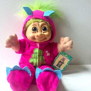 May include: A Russ Troll Kidz doll with bright green hair, wearing a hot pink dinosaur-themed onesie with blue felt spikes on the hood and feet, a green chest panel, and a small dinosaur graphic. A green product tag is attached to the doll's arm.