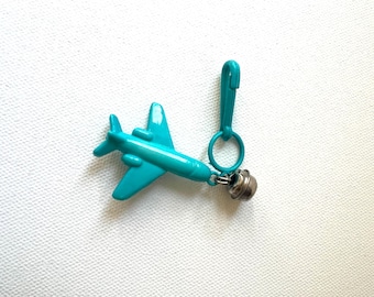 May include: A turquoise airplane charm with a small silver bell attached. The airplane is made of plastic and has a clip and ring for attachment. The charm is on a white textured background.