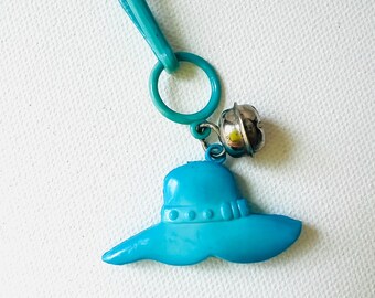 May include: A turquoise keyring with a sun hat charm and a small silver bell. The hat charm is made of solid, molded plastic with a wide brim and a detailed crown. The keyring includes a clasp and a ring for attaching keys.