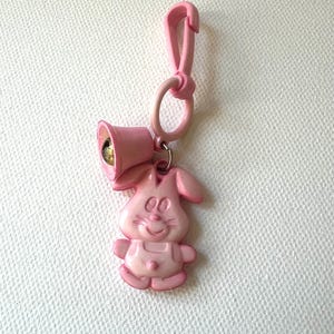 May include: A pink keychain featuring a cartoon bunny with a bell and a clip. The bunny has a smiling face and is wearing overalls. The bell is gold-colored inside. The clip and the ring are also pink.