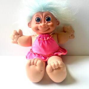 May include: A troll doll with light blue hair and large blue eyes. The doll has a peach-colored skin tone and is wearing a bright pink sleeveless dress with a ruffled hemline. The figure is in a seated position with arms extended outward.
