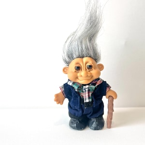 Könnte beinhalten: Eine Vintage-Troll-Puppe mit grauem Haar und einem karierten Hemd unter einer blauen Weste. Die Puppe hält einen Holzstock und trägt schwarze Schuhe. Ein klassisches Sammlerspielzeug.