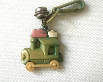 May include: A green and tan train keyring with a green clasp. The train features a pink and white cloud design. The keyring is made of plastic and has a silver-coloured bell attached.