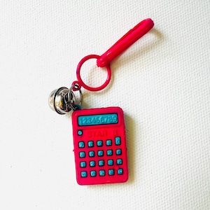 May include: A bright pink calculator keychain with a matching clasp and ring. The calculator has blue buttons and the text "STAR II" and numbers "123456789". A small silver bell hangs below the ring.