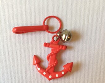 May include: A red anchor-shaped keyring with white polka dots and a silver bell. The anchor has a rope design and is attached to a red clasp and ring. Nautical-themed accessory.