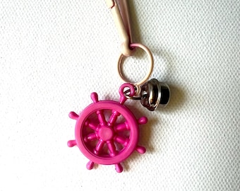 May include: A bright pink ship's wheel charm with a small silver bell and a pale pink clasp. The wheel is detailed and attached to a metal ring. The clasp is open, ready to attach to a keyring or bag.