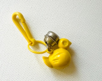 May include: A yellow duck-shaped keyring with a silver bell and a yellow clasp. The duck has a rounded body, a small beak, and a small head. The keyring is made of plastic and metal.