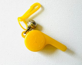 May include: A bright yellow whistle with a matching clip and a small silver bell attached. The whistle has a flower design on the front. The items are on a white background.