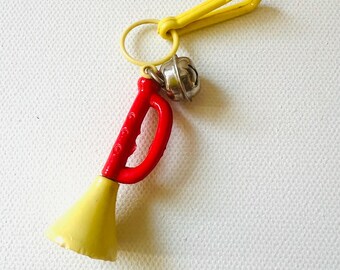 May include: A novelty keychain with a miniature trumpet. The trumpet has a red handle and yellow bell. A small silver bell and a yellow carabiner clip are attached. Suitable for keys or as a decorative accessory.