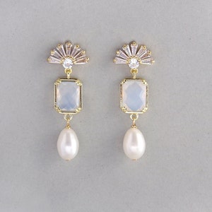 May include: Gold earrings with a white opal-like stone and a pearl drop. The earrings feature a fan-shaped design with clear rhinestones.