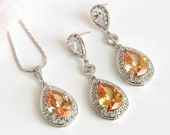 Puede incluir: Un collar con un colgante de lágrima perlada y pendientes a juego con una piedra grande, transparente, en forma de lágrima engastada en un marco de metal plateado. La piedra tiene un degradado de colores melocotón y naranja.