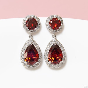 Könnte beinhalten: Ein Paar silberne Ohrringe mit einem tropfenförmigen roten Edelstein, der von kleineren klaren Edelsteinen umgeben ist. Die Ohrringe haben einen kleineren runden roten Edelstein oben.